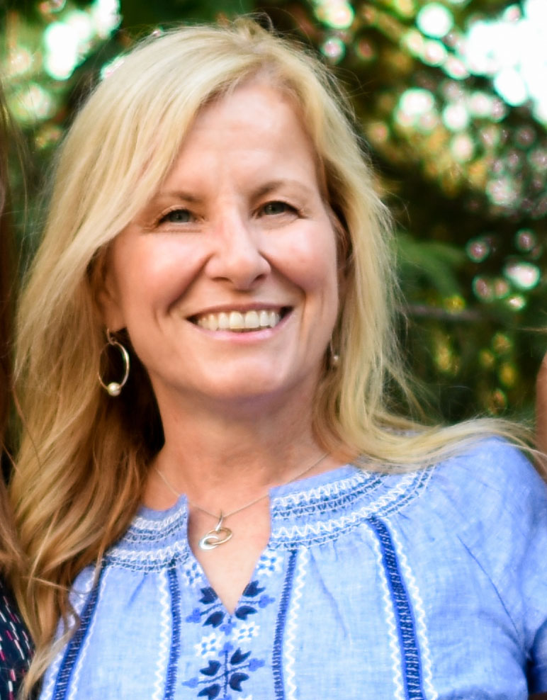 Smiling woman outdoors in a blue top with greenery in the background.